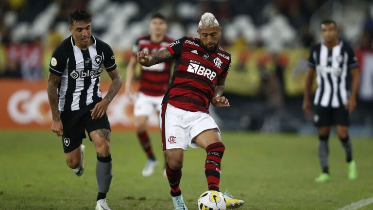 Jogadores de Flamengo e Botafogo disputam a posse de bola durante uma partida de futebol. O atleta do Flamengo, vestindo uniforme rubro-negro, conduz a bola enquanto é pressionado por um jogador do Botafogo, que veste uniforme listrado em preto e branco. O gramado apresenta marcas de desgaste, e ao fundo, torcedores acompanham o jogo nas arquibancadas.