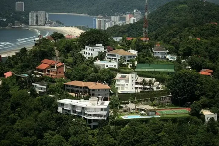 Vista aérea de uma área nobre em Santos, com várias mansões cercadas por vegetação, quadras esportivas e piscinas, tendo a praia e prédios ao fundo.