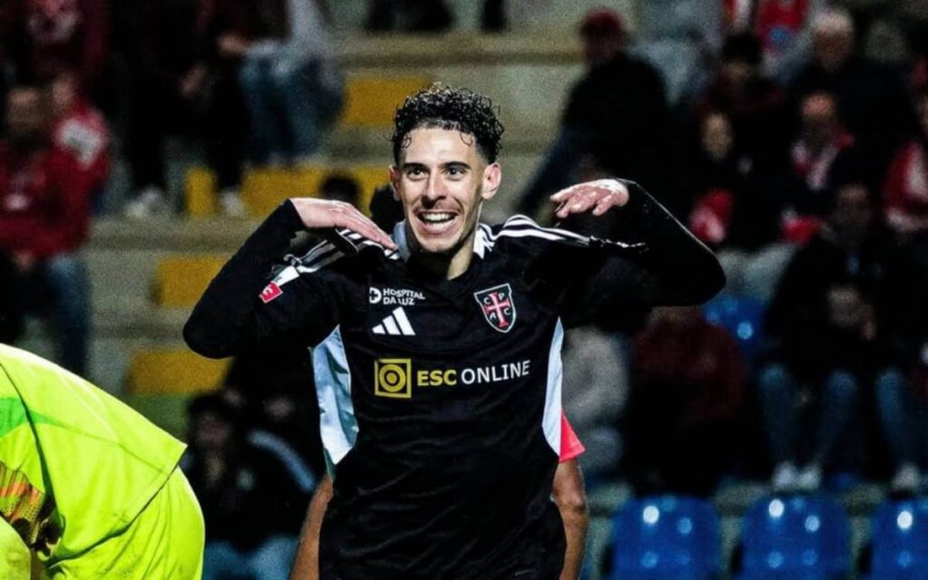 Jogador de futebol com uniforme preto do Casa Pia comemora um gol com um gesto característico, sorrindo e olhando para a câmera. Ao fundo, torcedores assistem à partida nas arquibancadas do estádio.