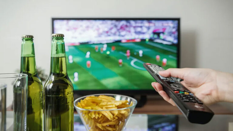 Uma pessoa assistindo a uma partida de futebol na TV, segurando o controle remoto, com duas garrafas de cerveja e uma tigela de batatas fritas sobre a mesa.