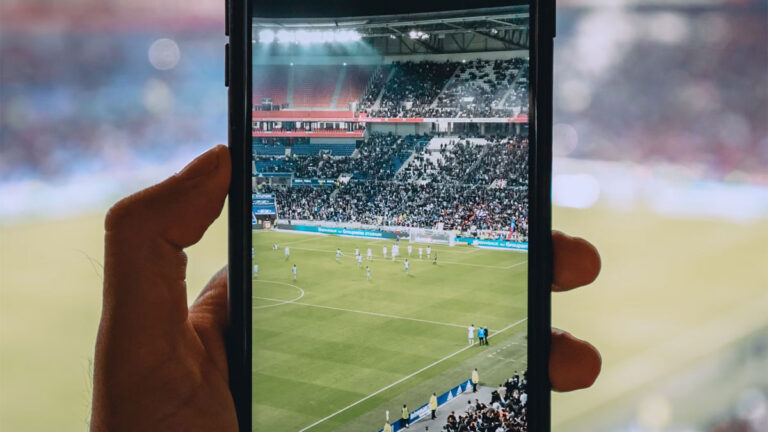 "Imagem de um estádio de futebol iluminado à noite, com um celular em primeiro plano gravando a partida em campo."